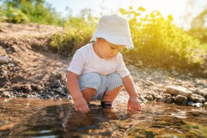 Playful little boy child playing with rocks and water by the river on a sunny day wearing hat in nature