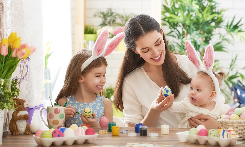 Celebrating Easter, a young girl and a baby wearing bunny ears a with an adult female helping them paint eggs